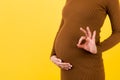 Cropped image of positive pregnant woman in brown dress showing okay gesture against her belly at yellow background. Easy and Royalty Free Stock Photo
