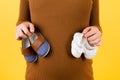 Cropped image of pregnant woman in brown dress holding two pairs of shoes for a boy and a girl at yellow background. Expecting Royalty Free Stock Photo