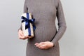 Cropped Image Of Pregnant Woman In Gray Dress Holding A Gift Box And Touching Her Belly At Gray Background. Expecting A Baby Boy.