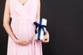 Cropped Image Of Pregnant Woman In Pink Dress Holding A Gift Box And Touching Her Belly At Black Background. Expecting A Baby Boy