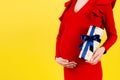 Cropped image of pregnant woman in red dress holding a gift box and touching her belly at yellow background. Expecting a baby boy Royalty Free Stock Photo