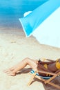 cropped image of woman in bikini laying on deck chair and holding cocktail in coconut shell under beach umbrella in front