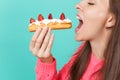 Cropped image of young woman in knitted pink sweater with closed eyes hold in hand, eating eclair cake isolated on blue