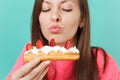 Cropped image of young woman in knitted pink sweater with closed eyes hold in hand, eating eclair cake isolated on blue