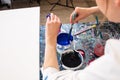 Cropped photo of hands of woman in white robe pouring paint from small plastic bottle into bucket with blue, mixing. Royalty Free Stock Photo