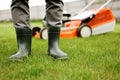 Cropped photo of male gardener legs in rubber boots stands on cut green grass lawn at backyard of house. Modern electric