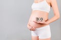 Cropped portrait of pregnant in white underwear woman holding cubes which show thirty weeks of pregnancy at gray background.