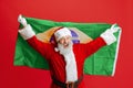 Cropped portrait of senior man wearing Santa Claus costume, holding Brazil flag isolated over red background. Football Royalty Free Stock Photo