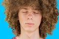 Cropped portrait of a young curly European man with long curly hair and closed eyes close up against blue background. very lush