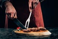 Chef in apron with meat fork and knife slicing gourmet grilled steaks with rosemary and chili pepper on wooden board Royalty Free Stock Photo