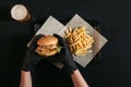 cropped shot of person in gloves holding tasty burger above tray with french fries and glass of beer on black Royalty Free Stock Photo