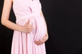 Cropped shot of positive pregnant woman in pink dress showing thumb up cool gesture over her baby bump at black background. Happy
