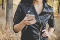 Cropped shot of slender female in black leather jacket holding smartphone at autumn forest background. Beautiful unrecognizable