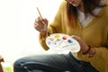 Cropped shot of young female artist is sitting on floor holding tube of oil paint and mixing colors on palette unfinished painting Royalty Free Stock Photo
