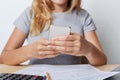 Cropped shot of young female enterpreneur in grey T-shirt, holding smart phone in hands, resting for minute after paper work at of Royalty Free Stock Photo