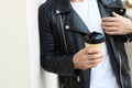 Cropped view of coffee to go in hand of blurred man in leather jacket near white wall Royalty Free Stock Photo