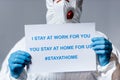 Cropped view of mature doctor in hazmat suit holding placard with i stay at work for you, you stay at home for us Royalty Free Stock Photo