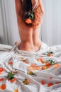 Cropped view of naked woman sitting backside on soft white bedding with orange red roses and petals and holding flower Royalty Free Stock Photo