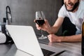 View of smiling man in medical mask holding glass of wine near laptop on kitchen worktop Royalty Free Stock Photo