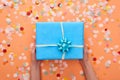 Cropped view of woman holding blue present with bow near confetti on orange. Royalty Free Stock Photo