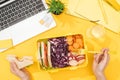 Cropped view of woman holding sandwich in hand near lunch box with food, laptop, glass of water and office supplies. Royalty Free Stock Photo