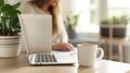 Cropped view of woman sitting at wooden table and working on laptop. A cup of coffee with a potted plant in the