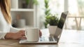 Cropped view of woman sitting at wooden table and working on laptop. A cup of coffee with a potted plant in the