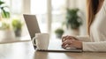 Cropped view of woman sitting at wooden table and working on laptop. A cup of coffee with a potted plant in the