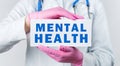 Cropped view of young female doctor in a white coat and pink sterile gloves holding a card with text MENTAL HEALTH