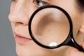 View of young woman with magnifier near problem skin on grey