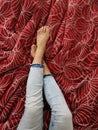 Crossed legs of a woman wearing light jeans on a red bed sheet with a floral pattern