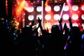 Crowd at a music concert, audience raising hands up in front of bright stage lights.