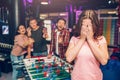 Cruel young people stand at table soccer in playing room and laughing. They point on woman in pink shirt. She cover face
