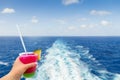 Cruise ship wake or trail on ocean surface with hand holding a glass with cocktail and straw