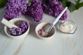 Crushed lilac petals Syringa vulgaris, yogurt and ready-made mask in a cup with a spatula close-up on a light marble background.