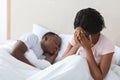 Crying african woman sitting in bed while her husband sleeping