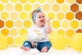 A crying dirty baby girl is sitting next to a broken cake. In the background is a pattern of yellow honeycombs and balloons. Smash