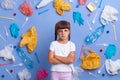 Almost crying little girl with pout lips wearing white t shirt posing against blue wall and much plastic garbage around, keeps Royalty Free Stock Photo