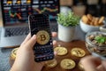 Cryptocurrency scene Hand holds Bitcoin, smartphone displays stock chart in cafe