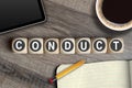 Cubes and dice on a wooden table with the word conduct with coffee, sketchbook and tablet computer