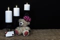 Cudlely teddy bear with pink bow on head, white candles perched on black candle holders on mesh place mat and wooden table with ca