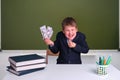 A cunning boy with money in his hands at a desk in a school classroom. US dollars in junior`s hand on a blackboard background, Royalty Free Stock Photo