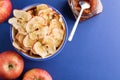 Cup with apple chips, fresh apples and glass jar of apple jam and metallic spoon on blue background
