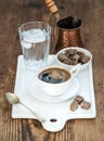 Cup of black coffee, copper pot, water with ice in glass and cane sugar cubes on white ceramic serving board over rustic