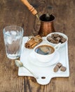Cup of black coffee, copper pot, water with ice in glass, cinnamon sticks and cane sugar cubes on white ceramic serving