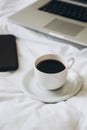 Cup of black coffee, laptop and smartphone on unmade white bed.