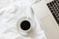 Cup of black coffee, laptop and smartphone on unmade white bed.