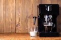 A cup of black coffee and a cup of milk stand near a coffee maker with a manual cappuccino maker. Wood background. Copy space for