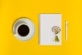 Cup of black coffee, notebook with blank paper, pen and bunch of daisy flowers on yellow background, top view