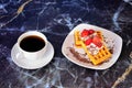 A cup of black coffee on a saucer and a plate with two waffles with strawberries, cream and chocolate edge on a marble table Royalty Free Stock Photo